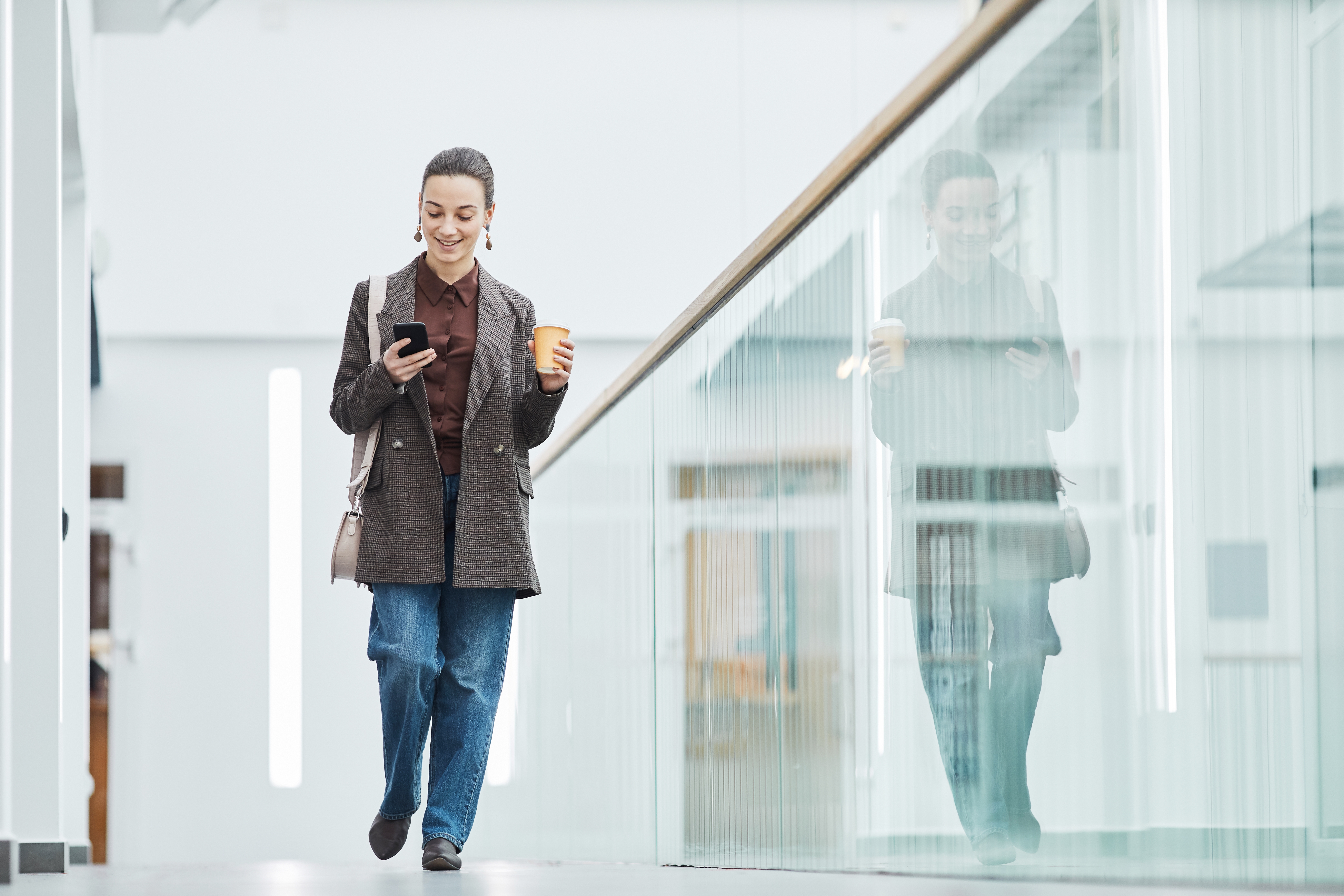 young gen z woman walks through large building looking at cell phone and drinking a coffee